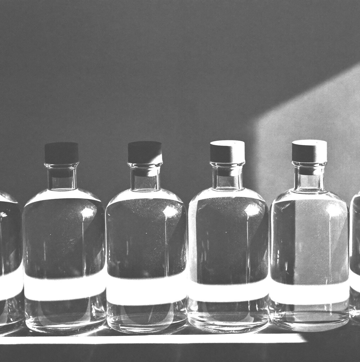 A row of identical unlabelled glass bottles on a shelf, hard side light, repeating shadows